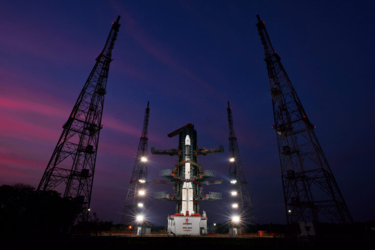 The image shows a large rocket on a launch pad at night. The rocket is surrounded by several tall towers with light shining on it. The sky behind the rocket is dark blue with some clouds.